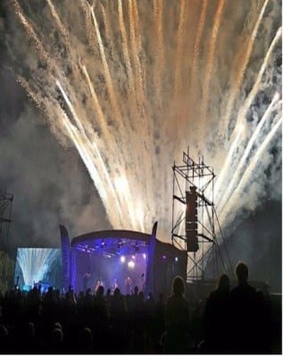 Fireworks erupting over a large outdoor concert stage at night
